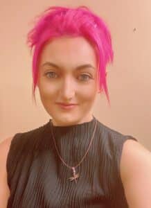 Gabrielle Rodriguez, a woman with light skin and pink hair, wearing a gray shirt and a dragonfly necklace.