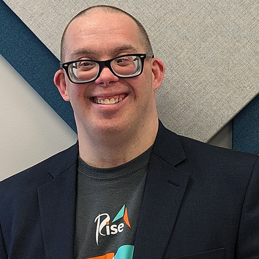 Jason Paddock smiles wearing a Rise t-shirt beneath a black suit jacket.