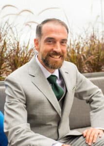 Dr. Brian Valentini, a white man with brown hair and a beard, wearing a grey suit with a green tie, poses for a professional headshot