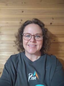 Beth Wilke, a white woman with curly brown hair and glasses, smiles for the camera. She is wearing a gray Rise shirt.