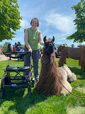 A person served by Rise stands next to a brown llama petting its fur with one hand. She smiles as she holds onto a walker with her other hand.