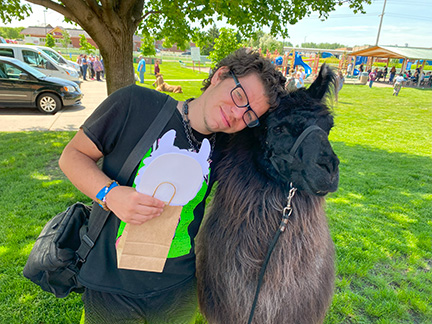 A person served by rise leans his head on a black and brown llama and smiles. The person and animal are standing in front of a park.