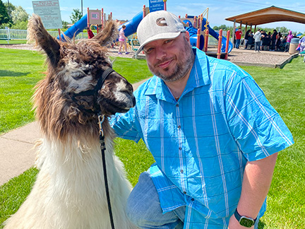 A man wearing a baseball cap and blue shirt sits on his knees while petting a white and brown llama.