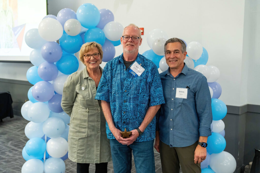 Tim Fahey accepts his award from Lynn Noren and Tim Dickie. There is a blue, purple, and white balloon arch behind them.