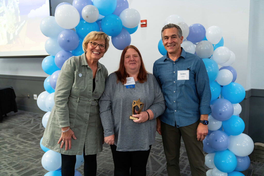 Lori Stellick-Wagner accepts her award from Lynn Noren and Tim Dickie. There is a blue, purple, and white balloon arch behind them.