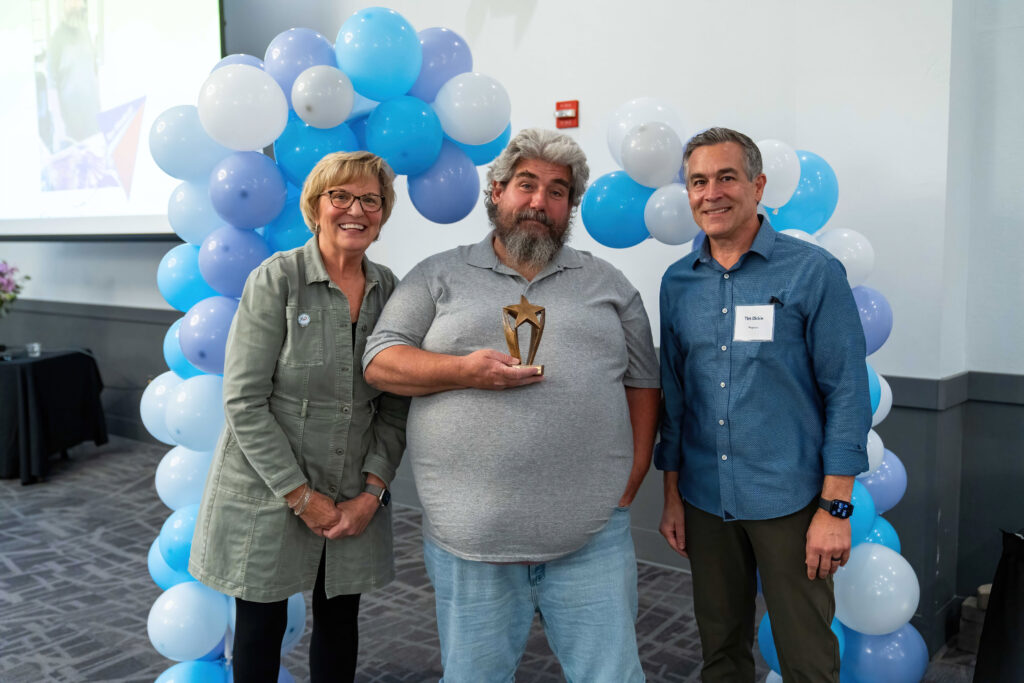 Ron Woolcott accepts his award from Lynn Noren and Tim Dickie. There is a blue, purple, and white balloon arch behind them.