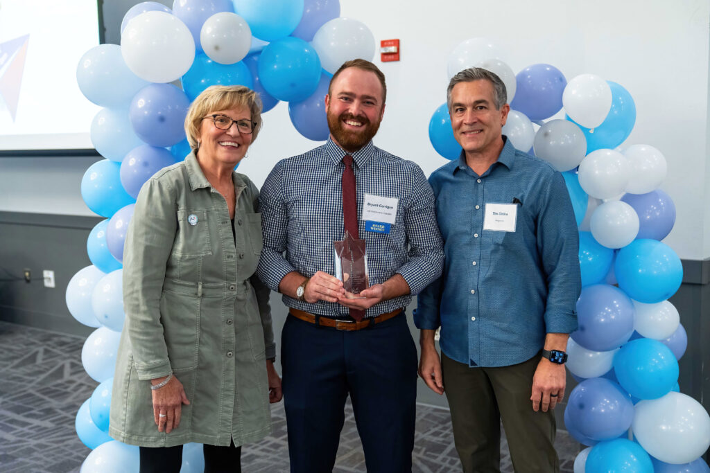 Bryant Corrigan accepts his award from Lynn Noren and Tim Dickie. They are framed by a blue, white, and purple balloon arch.