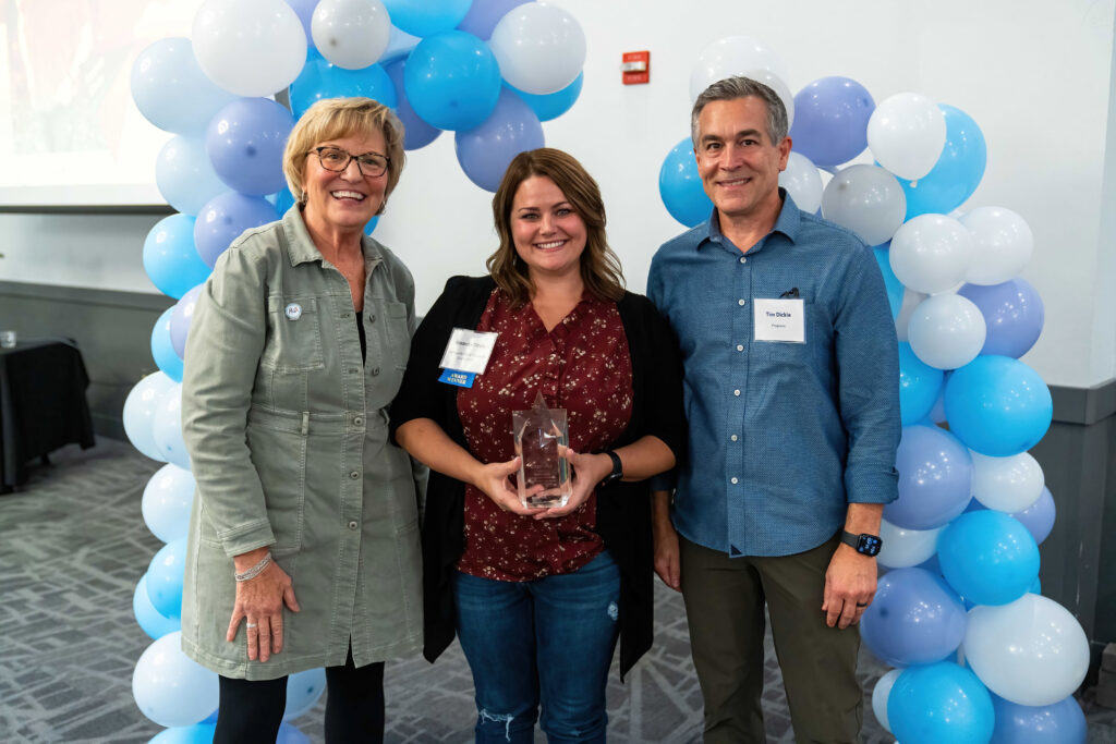 Amanda Dircks accepts her award from Lynn Noren and Tim Dickie. They are framed by a blue, white, and purple balloon arch.