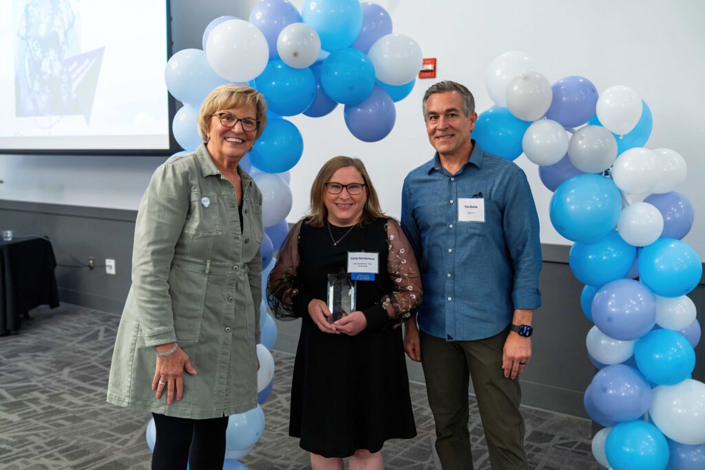 Katie McNamara accepts her award from Lynn Noren and Tim Dickie. They are framed by a blue, white, and purple balloon arch.