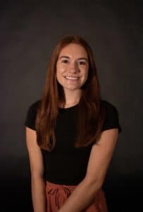 Woman posed for headshot in front of dark background, with long brown hair, black t-shirt, and smiling