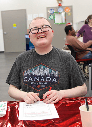 Person served by Rise wearing a grey t-shirt and smiling at the camera as they sit at the table and write on a piece of paper.