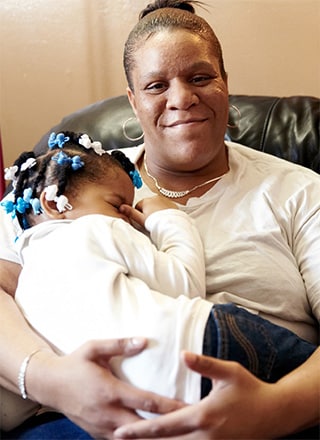 A mother sitting in a chair and holding her daughter while smiling at the camera.