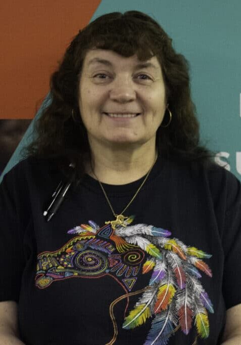 Janis Hooker smiles in front of a Rise branded wall wearing a black t-shirt with a colorful horse on it.