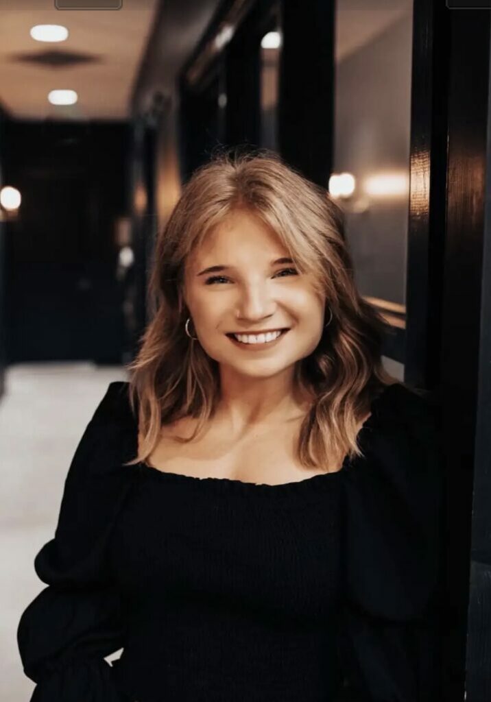 Champion of the Month, Olivia Booth, smiles in front of a dark wall.