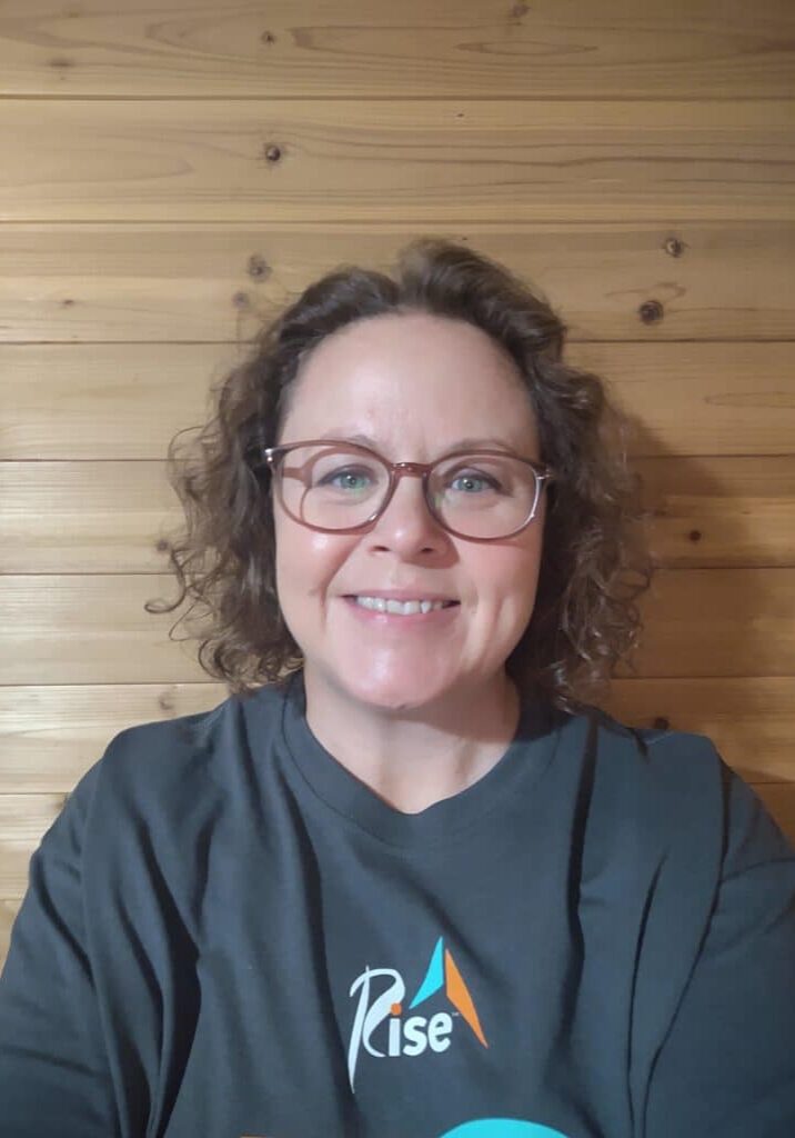 Beth Wilke, a white woman with curly brown hair and glasses, smiles for the camera. She is wearing a gray Rise shirt.