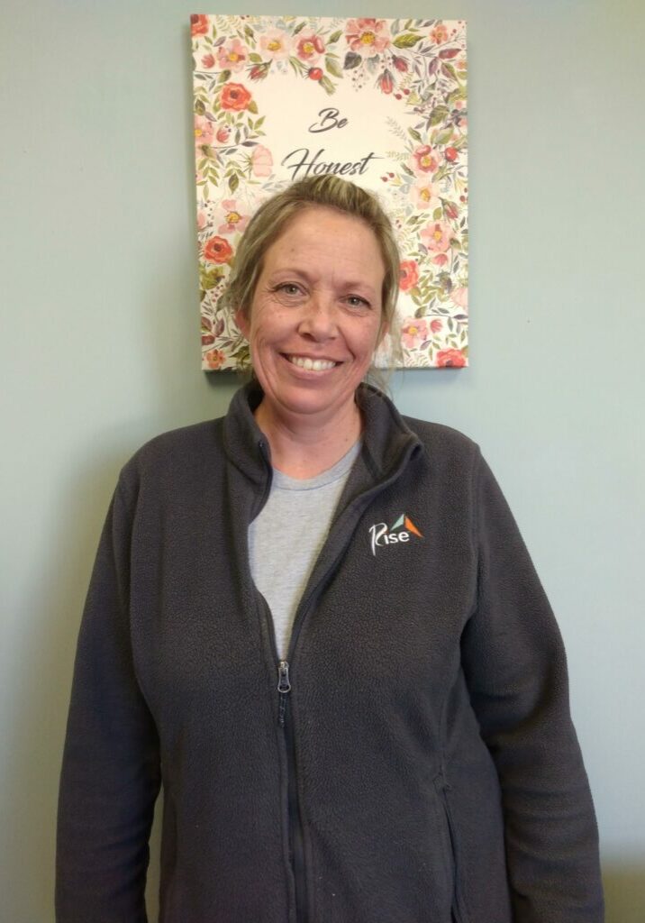 Jackie Gadach, a white woman with blonde hair, wearing a dark gray Rise branded zip-up over a light gray t-shirt, smiles for the camera. Behind her head is a piece of decorative art with flowers and an unlegible phrase.