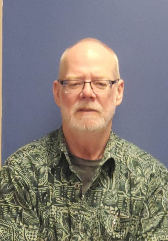 Tim Fahey, a lead DSP at Rise Crystal, poses in front of a blue wall. He wears a green printed shirt and glasses.