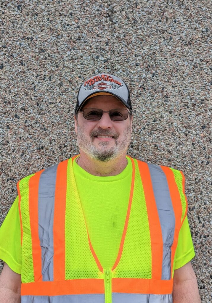 Champion of the Month John Barber smiles in front of a rock textured wall wearing a neon yellow and orange construction vest.