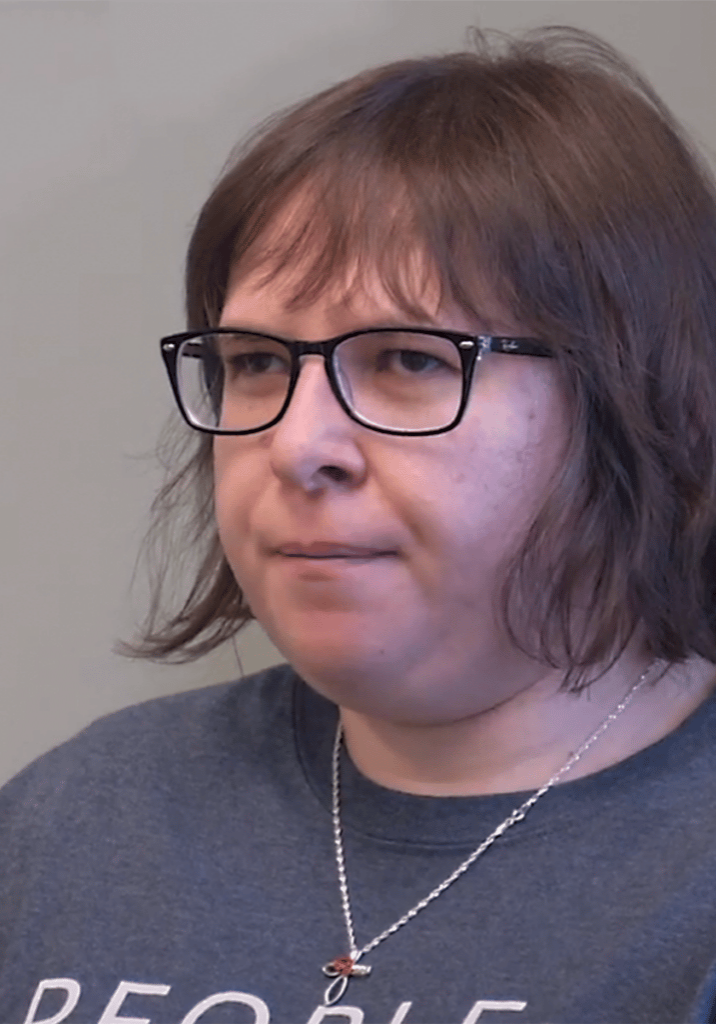 Amanda Lorenz sits in front of a gray background wearing black glasses, a gray shirt, and a silver necklace.