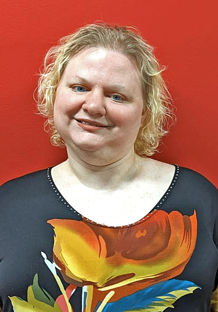 Amy Pancake smiles wearing a floral top with a red background behind her.