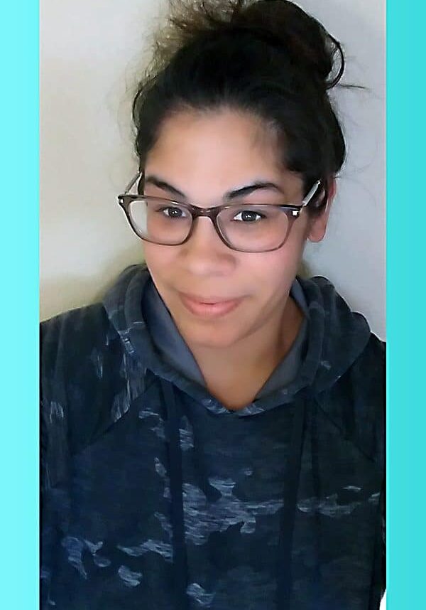 Andrea Myers smiles in front of a gray wall wearing glasses and a camouflage sweatshirt.