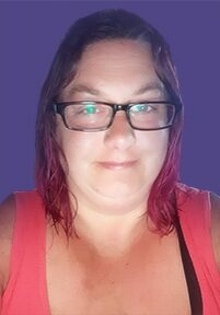 Angie Quitter smiles in front of a purple background wearing a pink tank top. She has black glasses and pink hair.