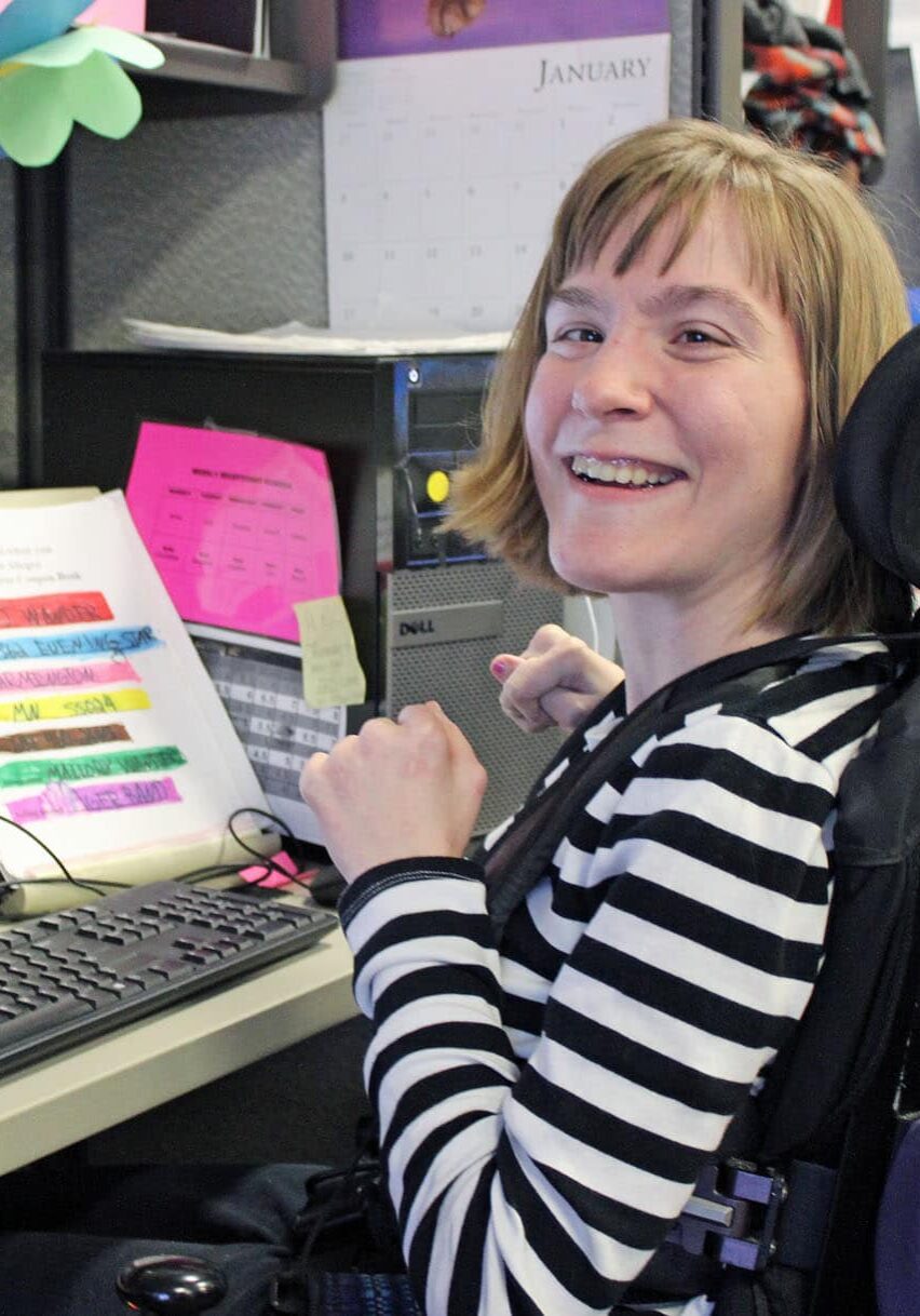 A woman with blonde hair wearing a black and white striped shirt sits at her desk in a wheelchair, smiling for the camera.