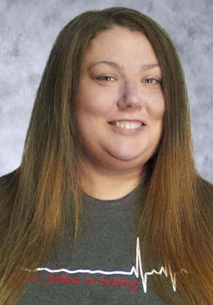 Ashley Wright smiles in front of a gray portrait background. She has long, straight brown hair and is wearing a gray t-shirt.