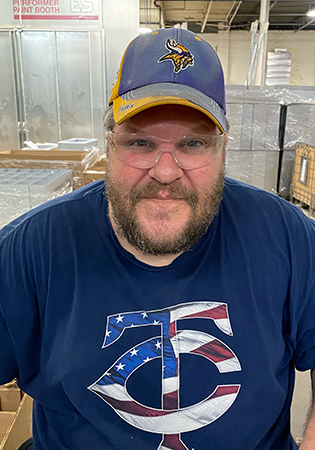 Brian Henley smiles in a warehouse wearing a Minnesota Vikings hat and Minnesota Twins t-shirt.