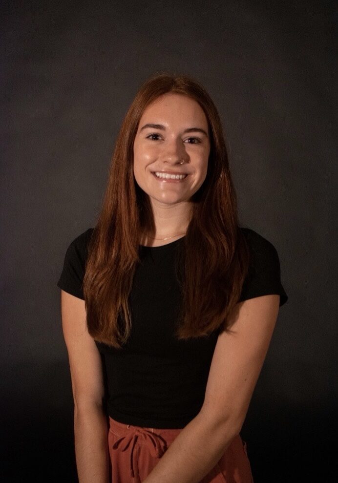 Woman posed for headshot in front of dark background, with long brown hair, black t-shirt, and smiling
