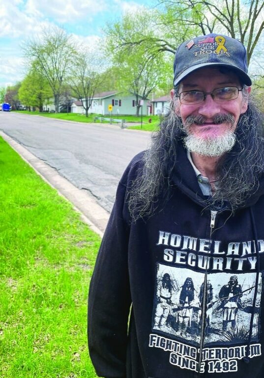 Cheyenne Johnson stands near grass and a sidewalk smiling wearing a black sweatshirt and baseball hat.