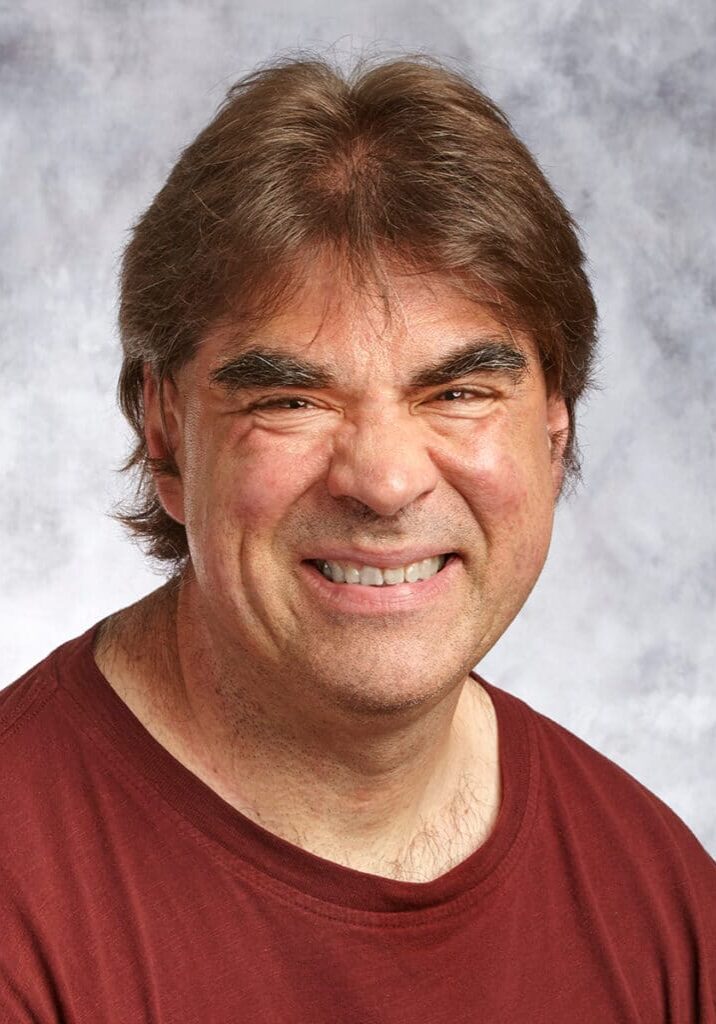 Chris Krause smiles wearing a maroon t-shirt in front of a gray portrait background.