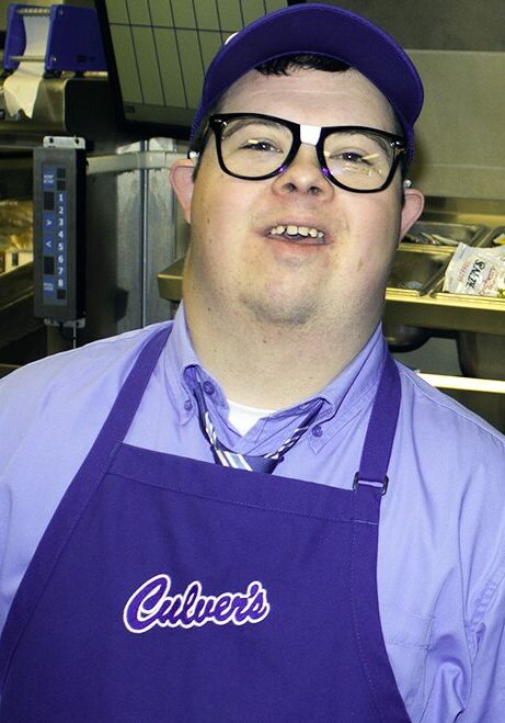 Wes Dahle smiles at his workplace, Culvers, wearing black glasses, a Culvers baseball hat, and a blue Culvers apron.