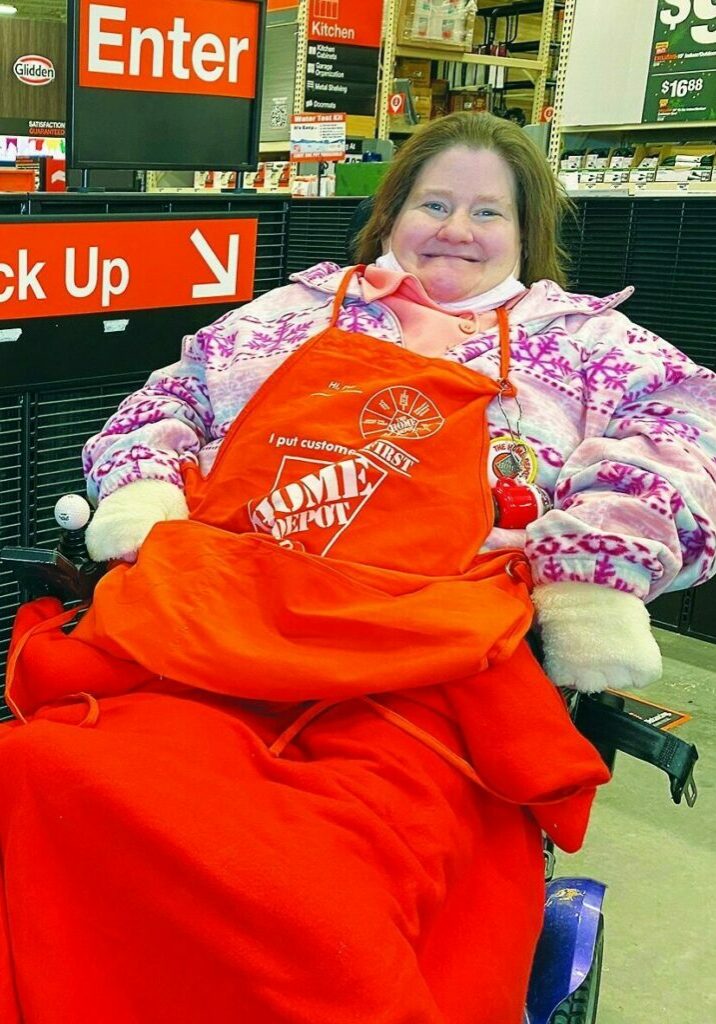 Denise Coppess sits in a wheelchair smiling as she greets people during her job at The Home Depot wearing an orange Home Depot apron.