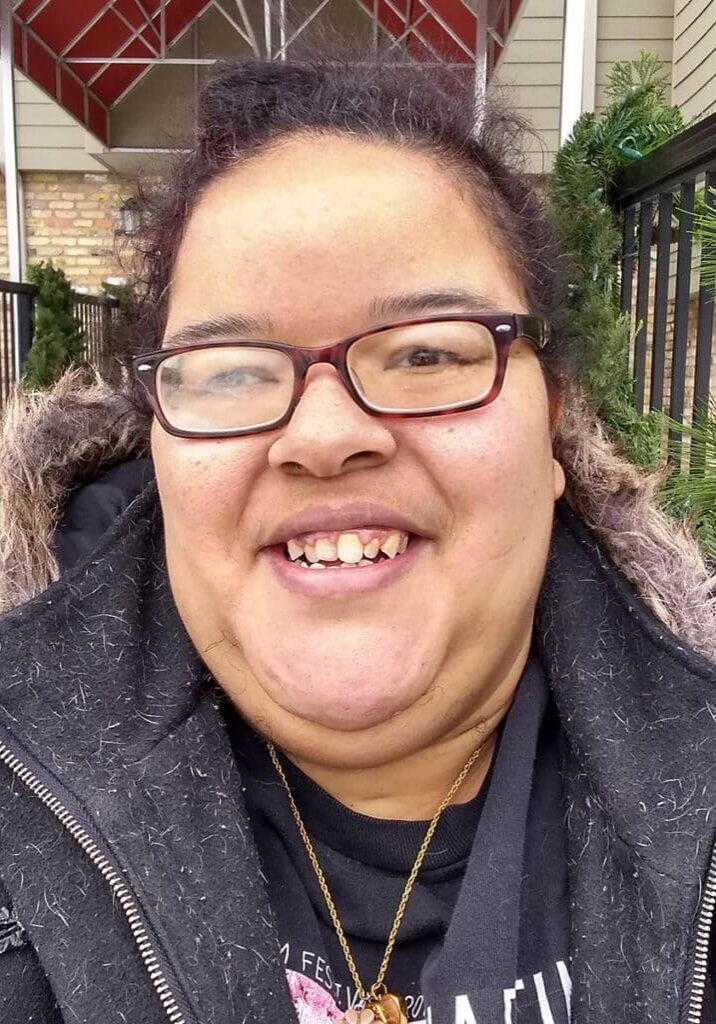 Elisa Raymond smiles in front of a building entryway wearing a black jacket and glasses.