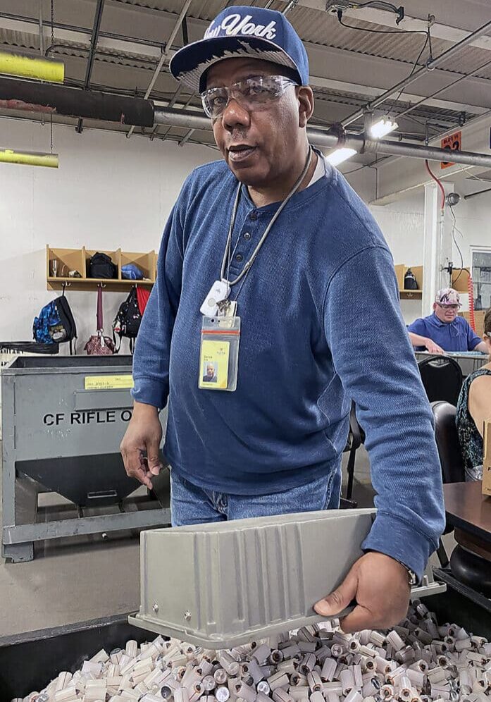 Derrick Smith pours a plastic tub of small, grey slugs into a large tub. Smith is wearing a blue sweatshirt and baseball hat as he smiles and looks at the camera.