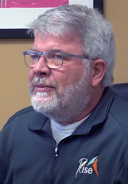 Mike Harper has a gray beard and wears a Rise zip-up and glasses while softly smiling.