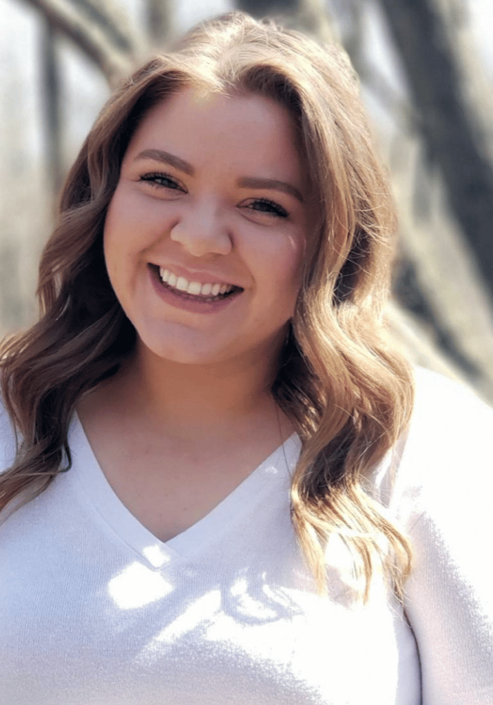 Hollie Peterson headshot smiling wearing a white long sleeve top while standing in front of a tree.