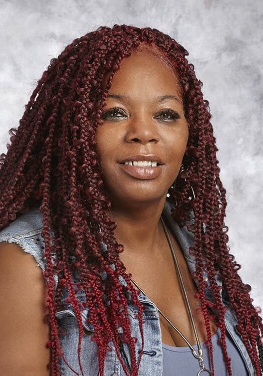 April Holman smiles in front of a gray portrait background wearing a denim vest with a blue tank top.