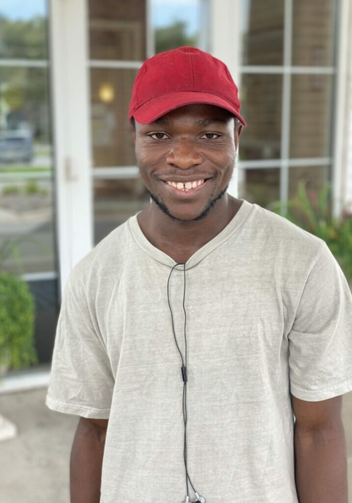 Jean Achida smiles outside wearing a tan t-shirt and a red baseball hat.