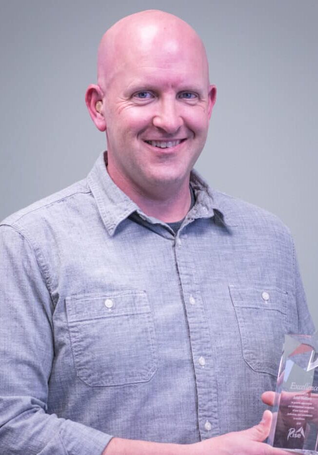 John Watters smiles wearing a gray button-up shirt while he holds a Rise award.