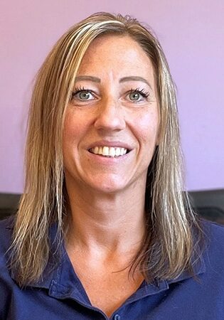 Kari Plautz smiles in front of a pink and purple wall wearing a blue blouse.