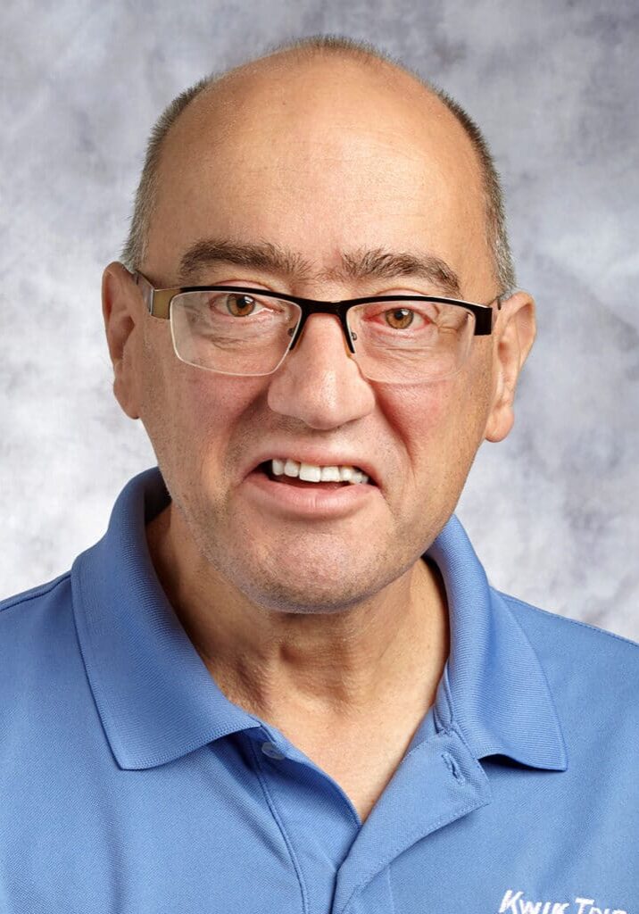 Kevin Symanitz smiles in front of a gray portrait wall wearing a light blue Kwik Trip work polo and glasses.