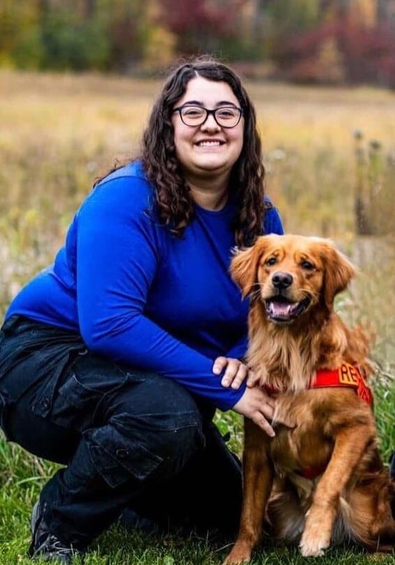 woman in blue shirt and glasses, crouching down to pose with golden retriever