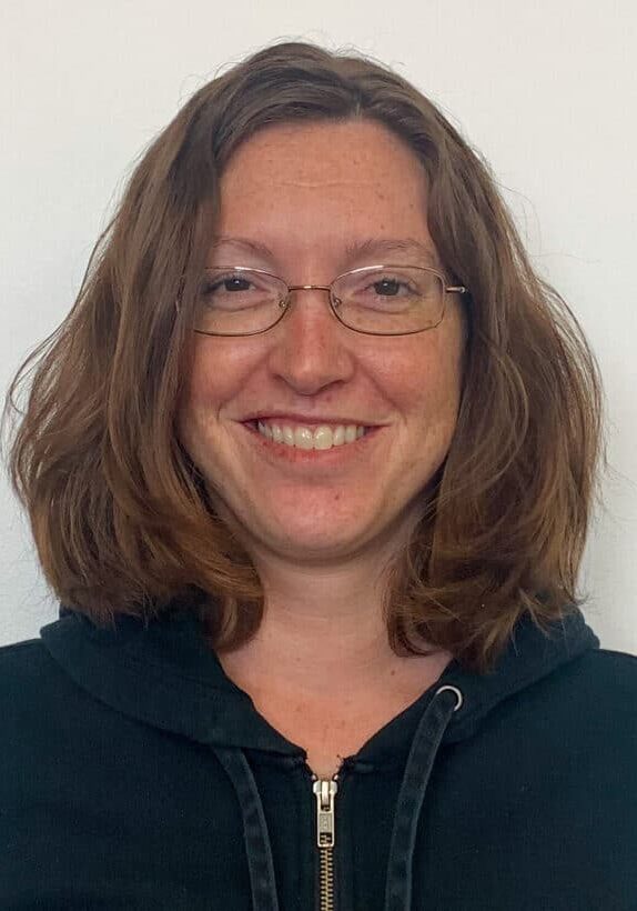 Lisa Bauman smiles wearing glasses and a black sweatshirt against a white wall.