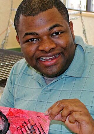 About Rise - Black man wearing blue shirt holds up a painting and smiles for the camera.