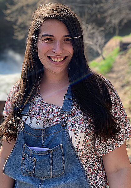 Mara Rosenberg smiles in front of a waterfall wearing overalls and a floral shirt.