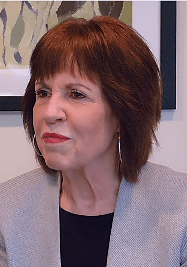 Mary Stransky soft smiling in front of artwork framed on a wall wearing a black top and gray blazer.