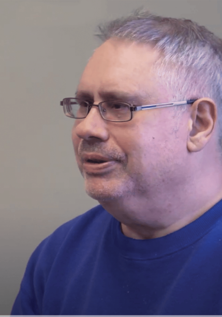 Paul Belair sits in front of a gray background wearing small glasses and a blue t-shirt.