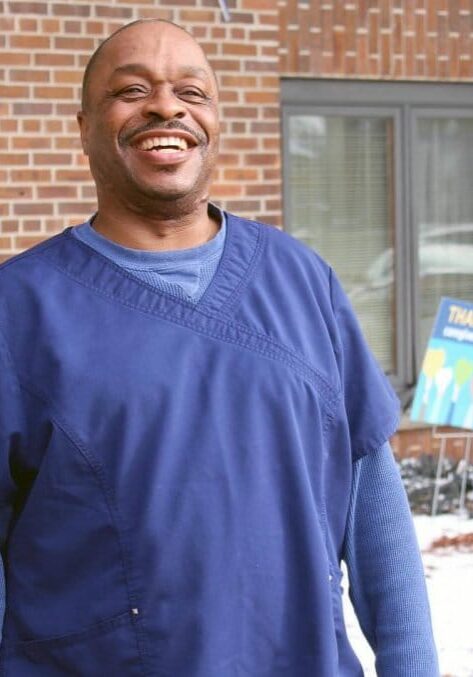 Ray Joyce smiles in front of a brick building wearing blue scrubs.